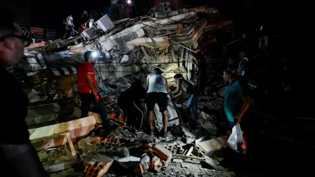 People inspect rubble of the aftermath of an earthquake, with recovery teams wearing headtorches.