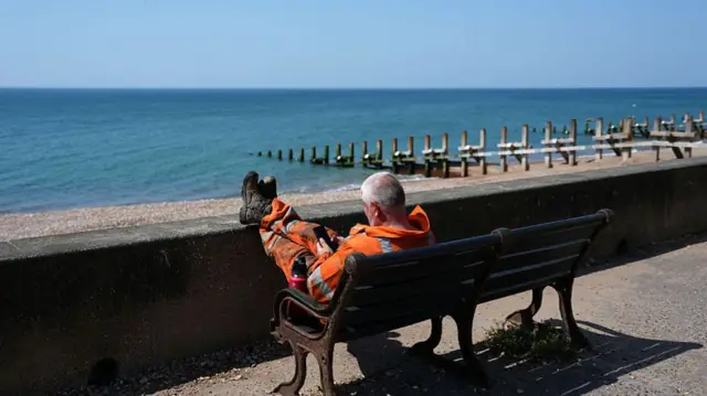 A worker takes a break from working in the heat sat on a bench with feet up, near a beach and the sea.