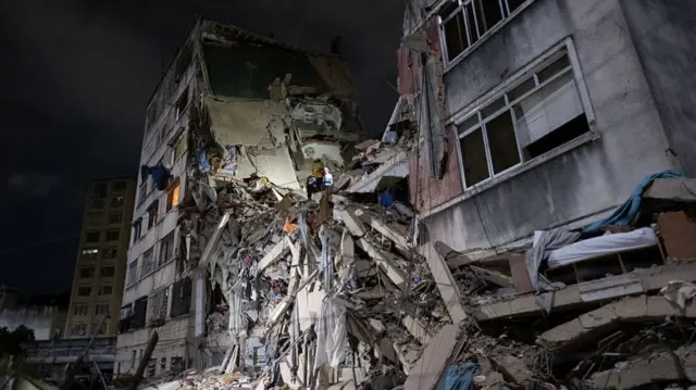 Rescue teams continue searching the rubble of a collapsed building at night.