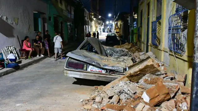 People gather on a street next to a crushed car