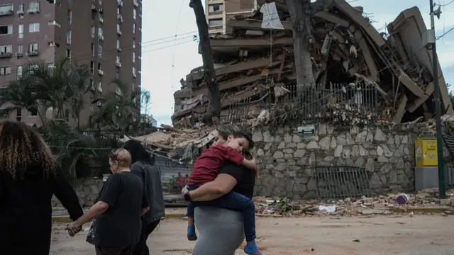 A woman in grey leggings and black t-shirt carries a young boy in a red shirt and blue trousers pajama, his head leaning on her shoulder. Behind him is a partially collapsed building