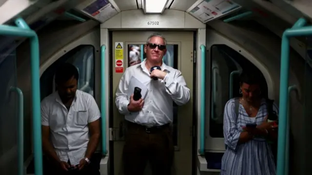 A man wearing a white shirt, black trousers and suglasses fans himself while standing in the middle of a tube carriage. Two other people stand to his right and left and are looking down at their phones.