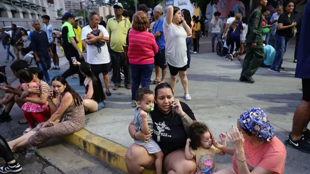 Multiple people standing and sitting at a kerb on a street in various brightly coloured clothes.