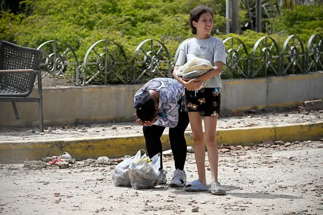 A woman bent over holds her head into her hands, two plastic shopping bags containing personal belongings at her feet. A young woman stands next to her in shorts and a grey t-shirt, holding a bag