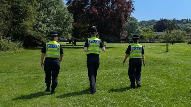 Three Wiltshire Police officers walking through a green park on a sunny day.