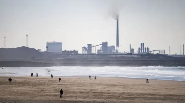 A large expanse of flat sand is shown on a beach, with people walking. There is a large industrial site in the background.