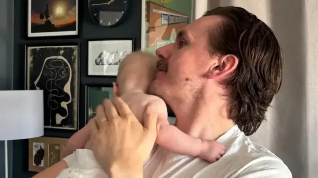 Jackson Bylett is pictured with his five-month-old baby who is wearing a nappy and facing away from the camera.