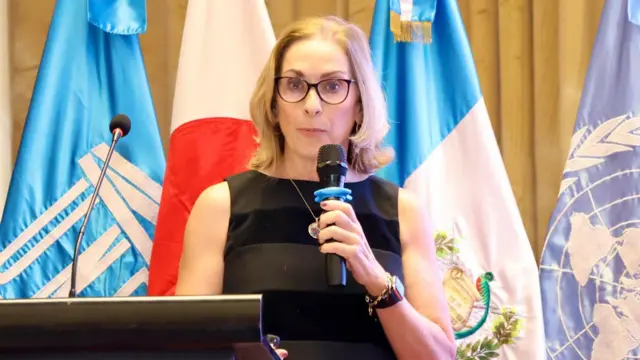 América Bendito speaking into microphone wearing black dress with flags behind her