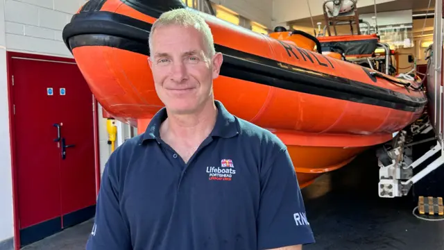 Chris Wade wearing a blue RNLI polo shirt, standing in front of an inflatable orange lifeboat dinghy.