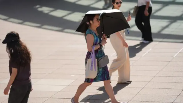 A woman walks across the frame as she uses an umbrella to shade herself from the sun