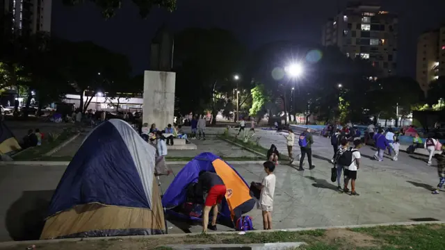 In the foreground is a set of tents with children moving things inside the smallest one. The scene appears to be set in a park in Caracas, with several other people with backpacks standing and walking around the background