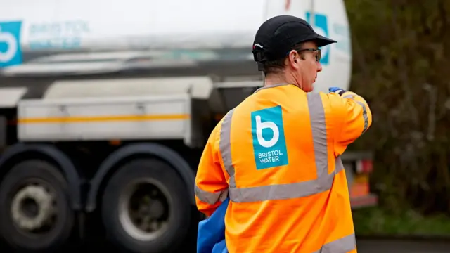 A Bristol Water workman wearing branded orange hi-vis overalls. In the background is a branded Bristol Water truck.