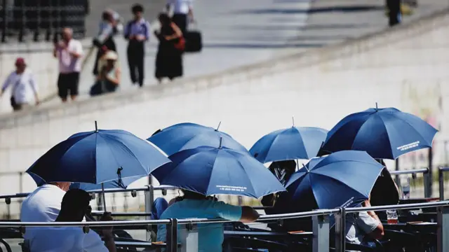 People on a boat sat under blue umbrellas, as people walk in the background.