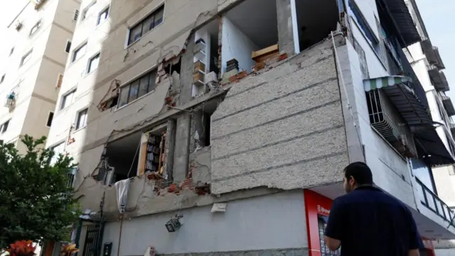 A man stands outside a damaged building, the inside is exposed by a hole in its side.