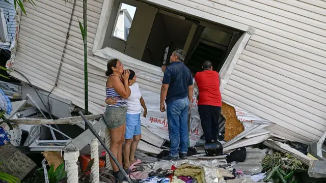 A woman looks at the remains of a white house with an anguished look, another young woman hugging her one-armed as two men look into the property