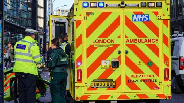 Two people in uniform stood by a London Ambulance van.
