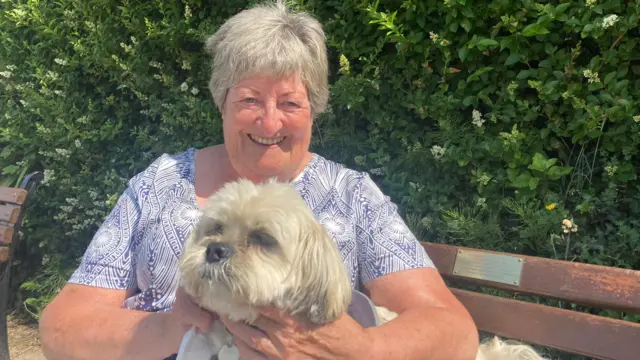 A woman and a small dog with white fur. The dog is sitting on the woman's knee. She's sitting on a bench. She has short hair and is wearing a blue and white pattered top. Bushes are in the background.
