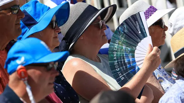 A woman in a crowd fans herself, she is surrounded by people wearing sunhats.