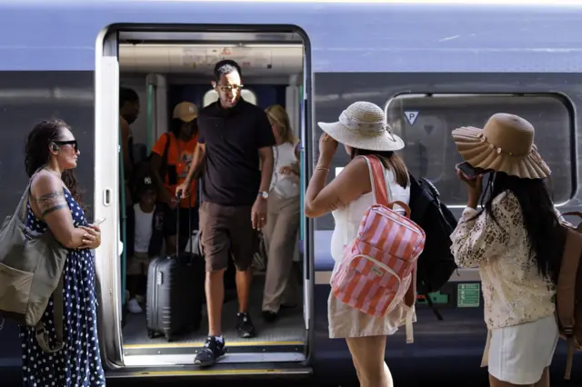 Passengers wearing hats wait to board a train as a passenger wearing shorts and a t-shirt gets off