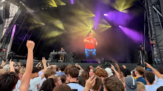 A crowd of people are dancing and looking towards a large state, with a DJ playing on turntables.