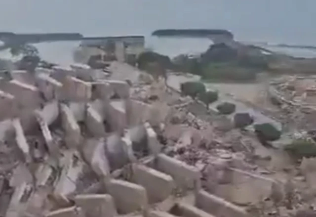 A screengrab from the verified video showing a collapsed multi‑storey building reduced to large slabs of debris beside a body of water under a hazy sky.