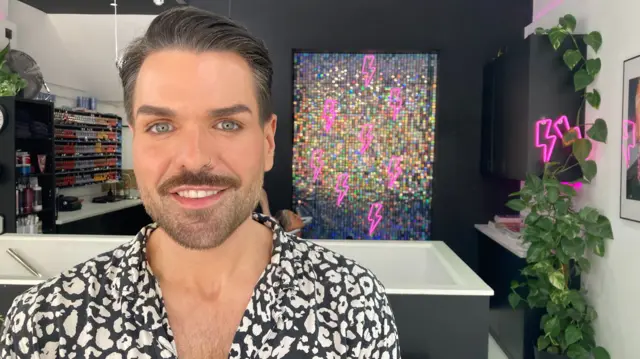 Feargal Doyle.wearing a black and white leopard print shirt and a silver nose ring. He has light blue eyes and dark hair, styled neatly with a trimmed beard. He is standing in his hair salon, with a holographic disc wall behind him, decorated with pink lightning bolts.