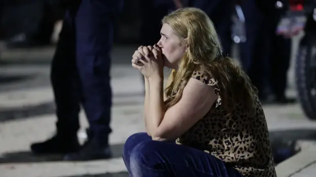 A woman clasps her hands together as she sits on the side of a road.