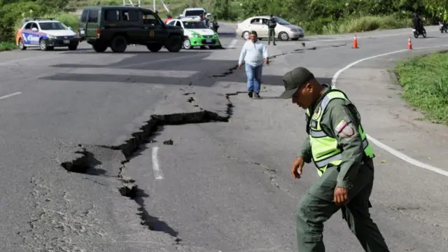 A member of the national guard wearing a green florescent vest and uniform steps over a large crack in the road