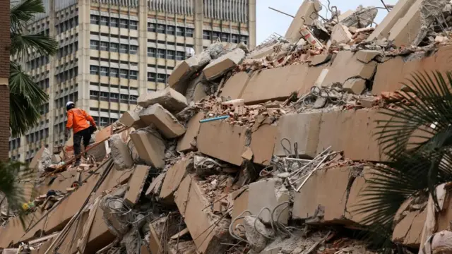 A rescue worker in a orange jacket walks over the remains of a building. Large slabs of concrete lie in between rubble. A second tower block can be seen standing in the background