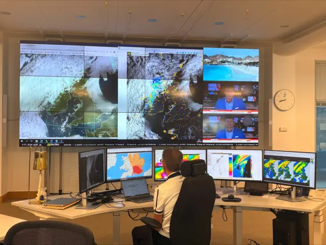 Forecaster sitting at desk in front of five small screens with a large one on the wall in front of them