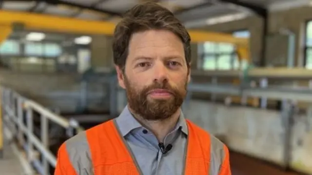 A man with a thick brown beard and brown hair wearing an orange high vis South East Water jacket. He is standing in a water treatment works site.