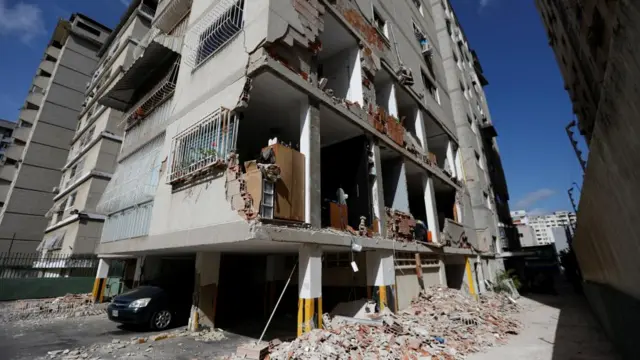 The side of a building has been reduced to rubble revealing the insides of four room. A pile of rubble can be seen at the edge of the building
