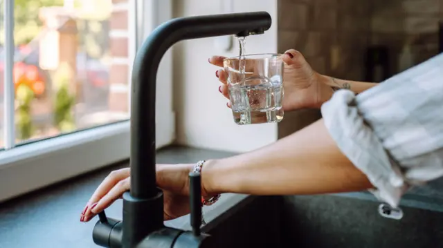 A stock image of a woman filling up a glass of water from a matt black kitchen tap.