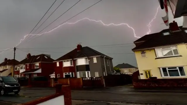 A bolt of purple lightening stretching across a dark sky above a row of semi-detached houses.
