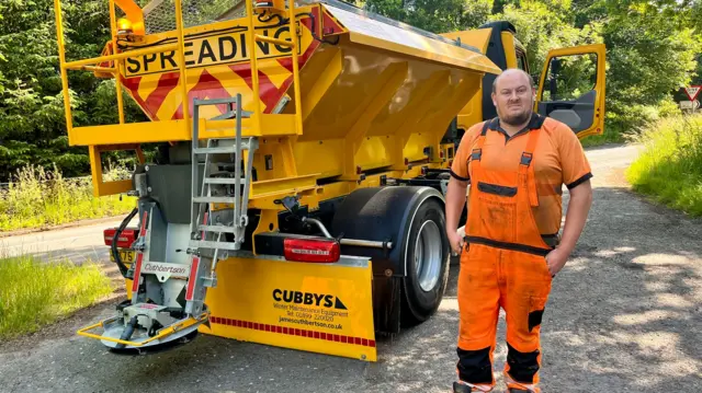 A man in orange overalls is pictured standing beside a yellow grit-spreading truck.