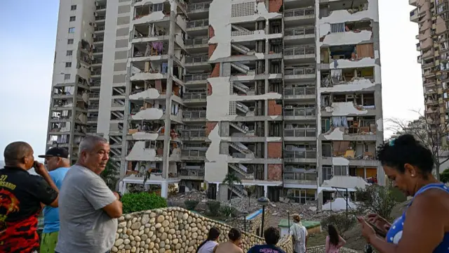 A crowd of people mill around on some steps looking onto an apartment building that is very damaged
