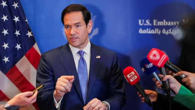 Marco Rubio speaks to reporters in front of a US flag. Several microphones can be seen close to Rubio