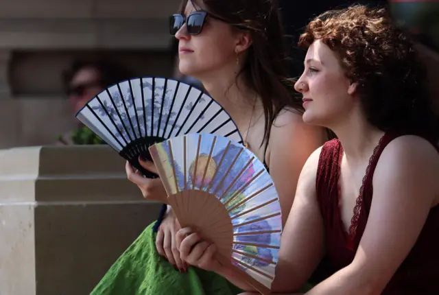 Two young women wearing summer clothing sit in the shade of building as they waft themselves with fans.