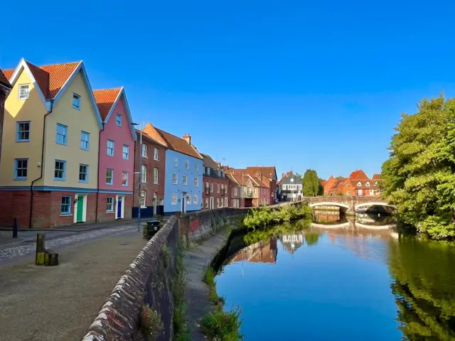 Sunny skies over a river, with picturesque brightly-coloured houses on one side of the riverbank
