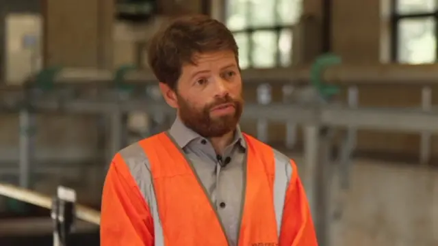 A man with short aubern hair and a beard wears a grey shirt and orange high vis jacket while standing in a water treatment plant