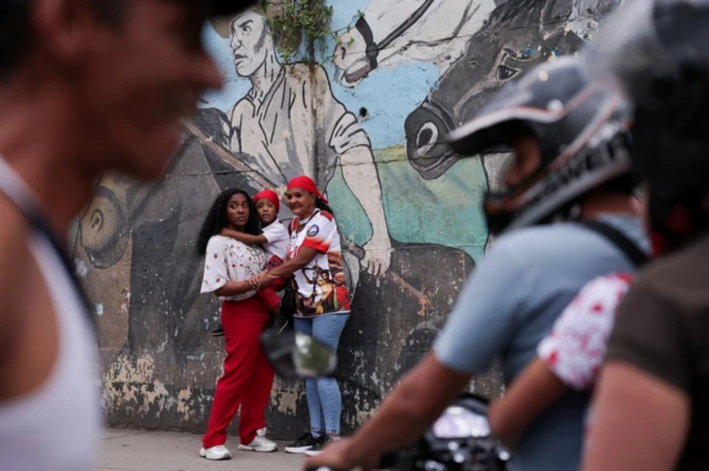 A young woman in red trousers holds a toddler, an elderly woman in a red cap with her arms around both of them. In the foreground are people walking by, and a man riding a bike, a young girl with her arms around him