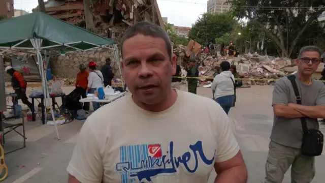 Erick Paul Martínez Santos looks to camera as he speaks - the rubble of a building can be seen behind him