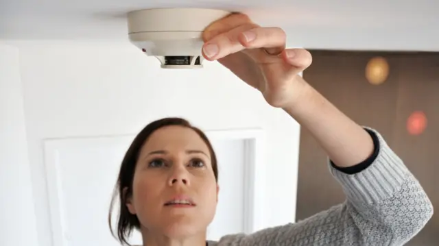 A woman with dark hair reaches up and touches a white fire alarm on a ceiling.
