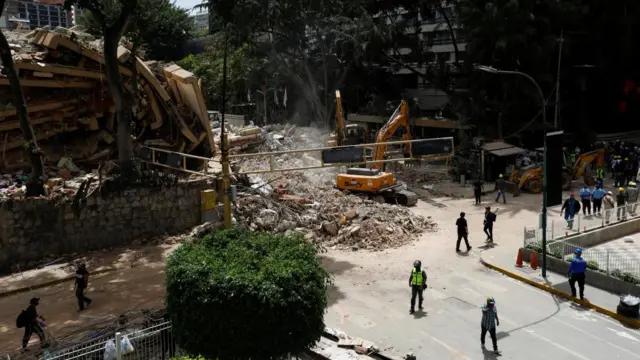 Two diggers work in a pile of rubble next to a collapsed building. Several workers are walking past the scene