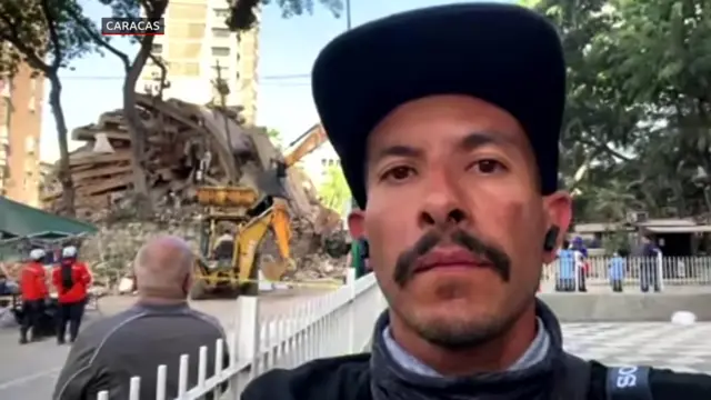 A journalist wearing a black baseball cap looks to his phone camera with a pile of rubble in the background.