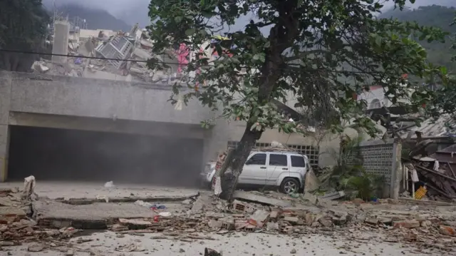 A silver car is seen among rubble, with a tree in front and more rubble in the background.