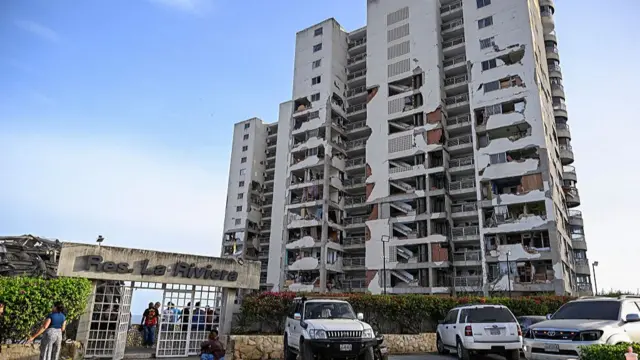 Wide short of a heavily damaged building. The entrance, a white gate off to the left, has a sign reading Res La Riviera on top. People are seen standing near the white entrance gate. Parts of the external walls have collapsed, showing the inside of flats