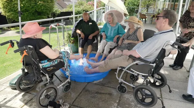 Five care home residents sitting on a patio terrace with their feet splashing in a blue plastic paddling pool.