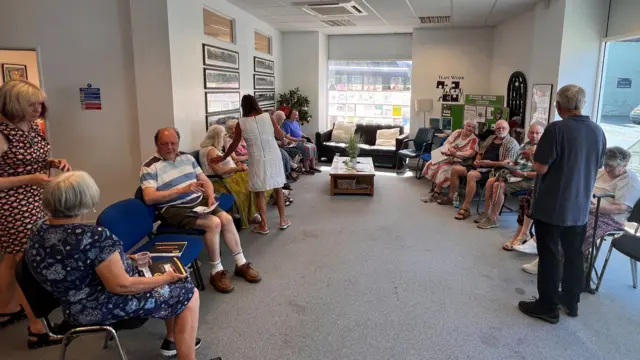 A group of people sit on chairs in a room - a couple of people are standing. It is a room that looks like it has been converted from an office.