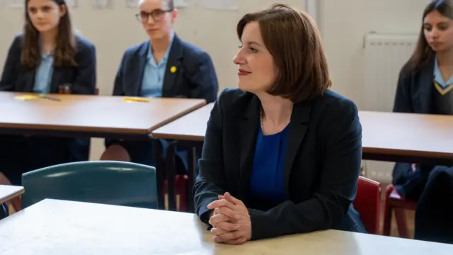 Bridget Philipson, the education secretary, during a visit to a school. She is sat at a school desk with some female pupils sat behind her.